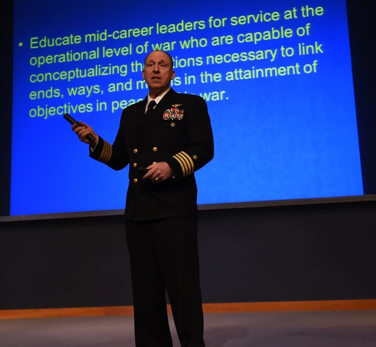 Military official speaking in front of a presentation screen showing transcription